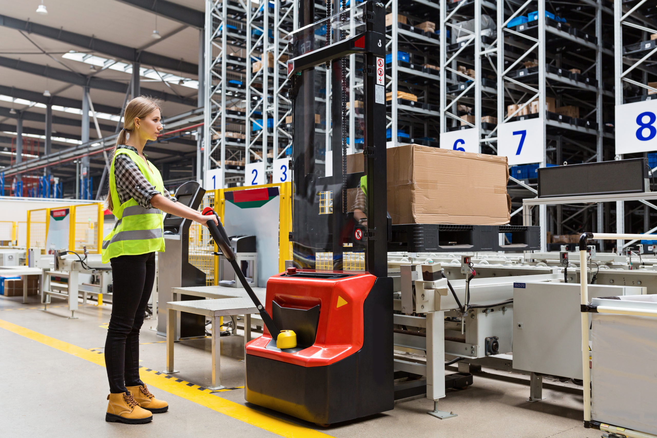 Storehouse woman employee in uniform using forklift with box in modern automatic warehouse.Boxes are on the shelves of the warehouse. Warehousing, machinery concept. Logistics in stock.